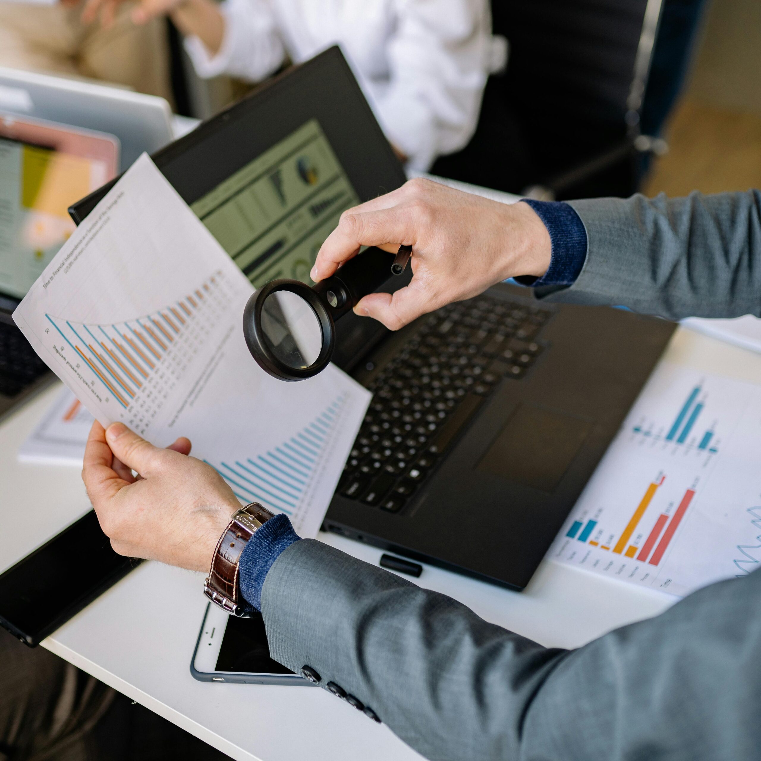 Businessman examines data charts with a magnifying glass over a laptop in an office setting.